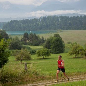 Laufen & Nordic Walking Mann wandert mit Stöcken auf einem Grasweg mit Bergen und Wald im Hintergrund
